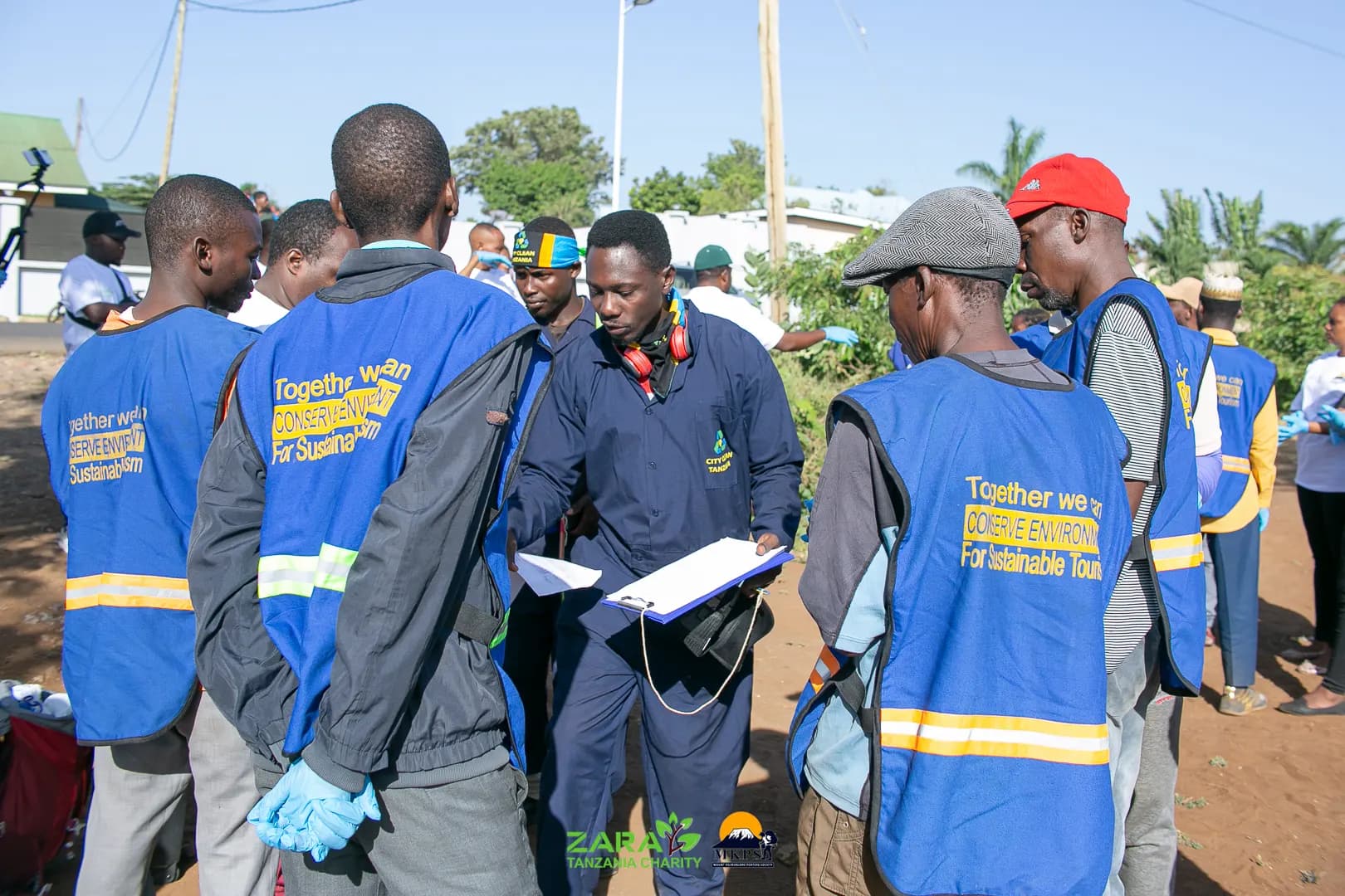 Cleanup volunteers in blue environmental vests gathered around a supervisor holding a clipboard at the start of a community cleanup day.