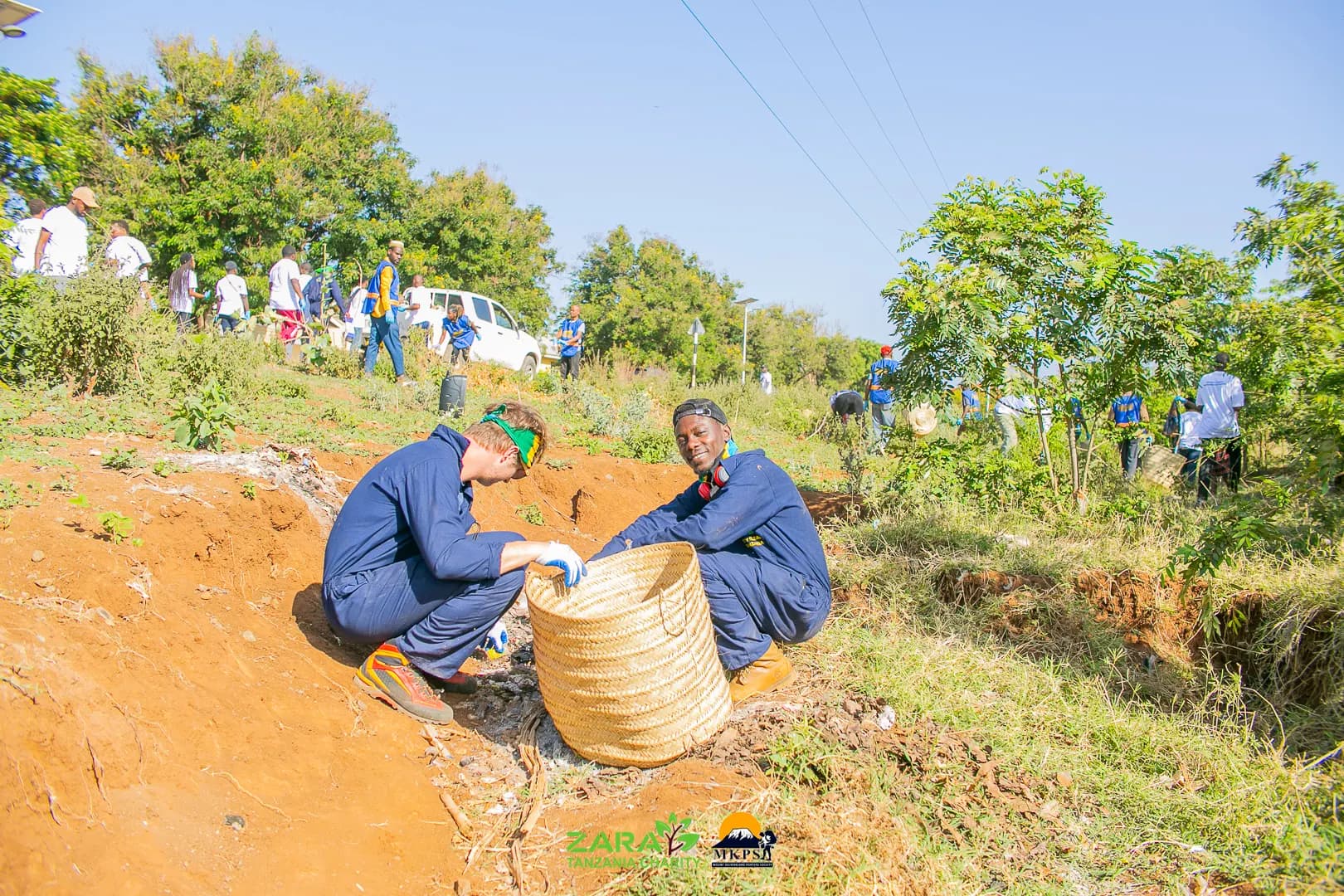 Two volunteers in blue overalls crouched on a bare earth bank, steadying a woven basket as they clear litter from the slope.