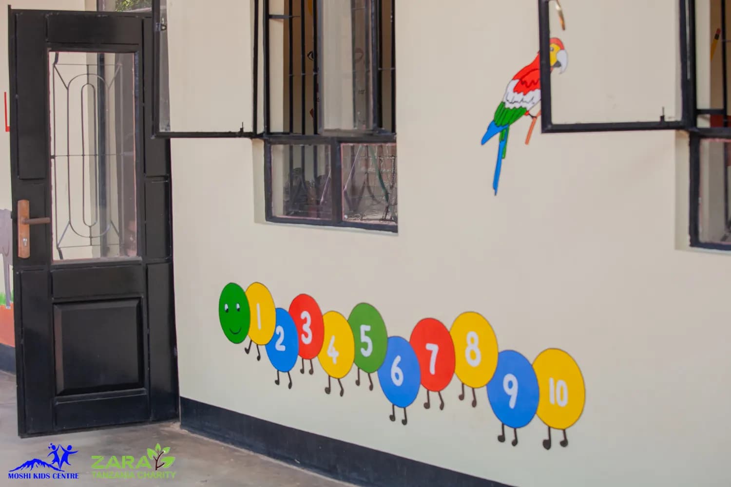 The painted exterior wall of Moshi Kids Centre, with a counting caterpillar numbered one to ten and a parrot painted beside the classroom door.
