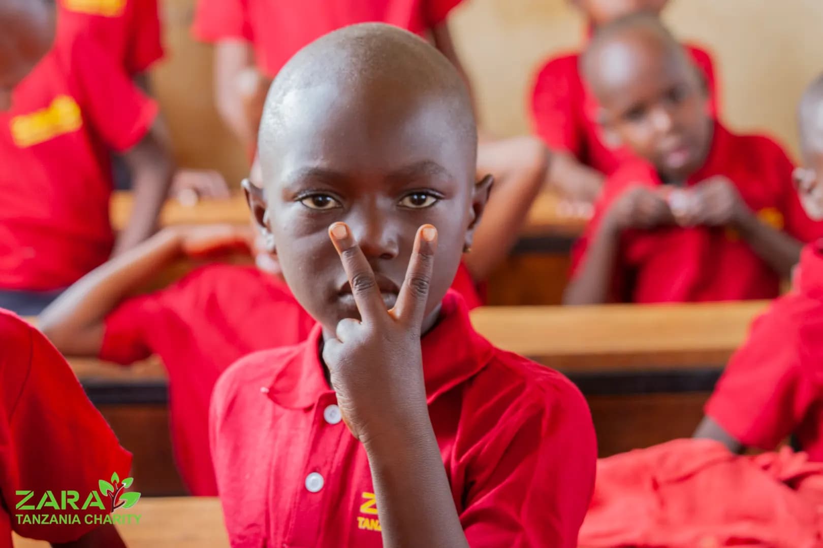 A boy in a red charity polo shirt sitting at his classroom desk, holding up two fingers and smiling at the camera.