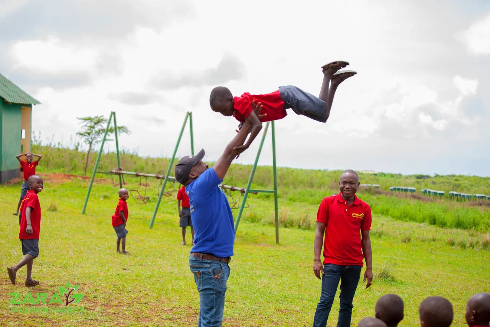A staff member lifting a small child high into the air beside the swings on the school field, other children and staff watching from the grass.