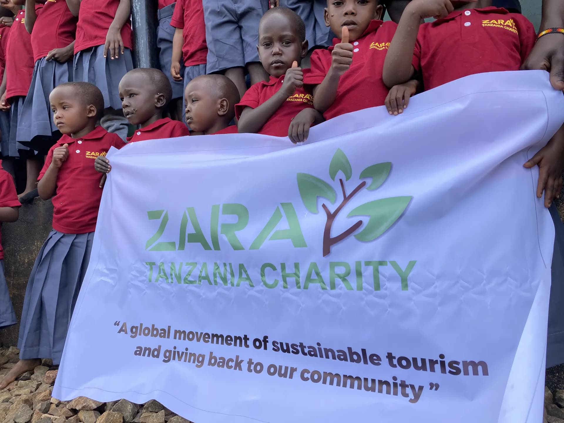 Pre-school children holding the charity banner, its motto — a global movement of sustainable tourism and giving back to our community — clearly readable.