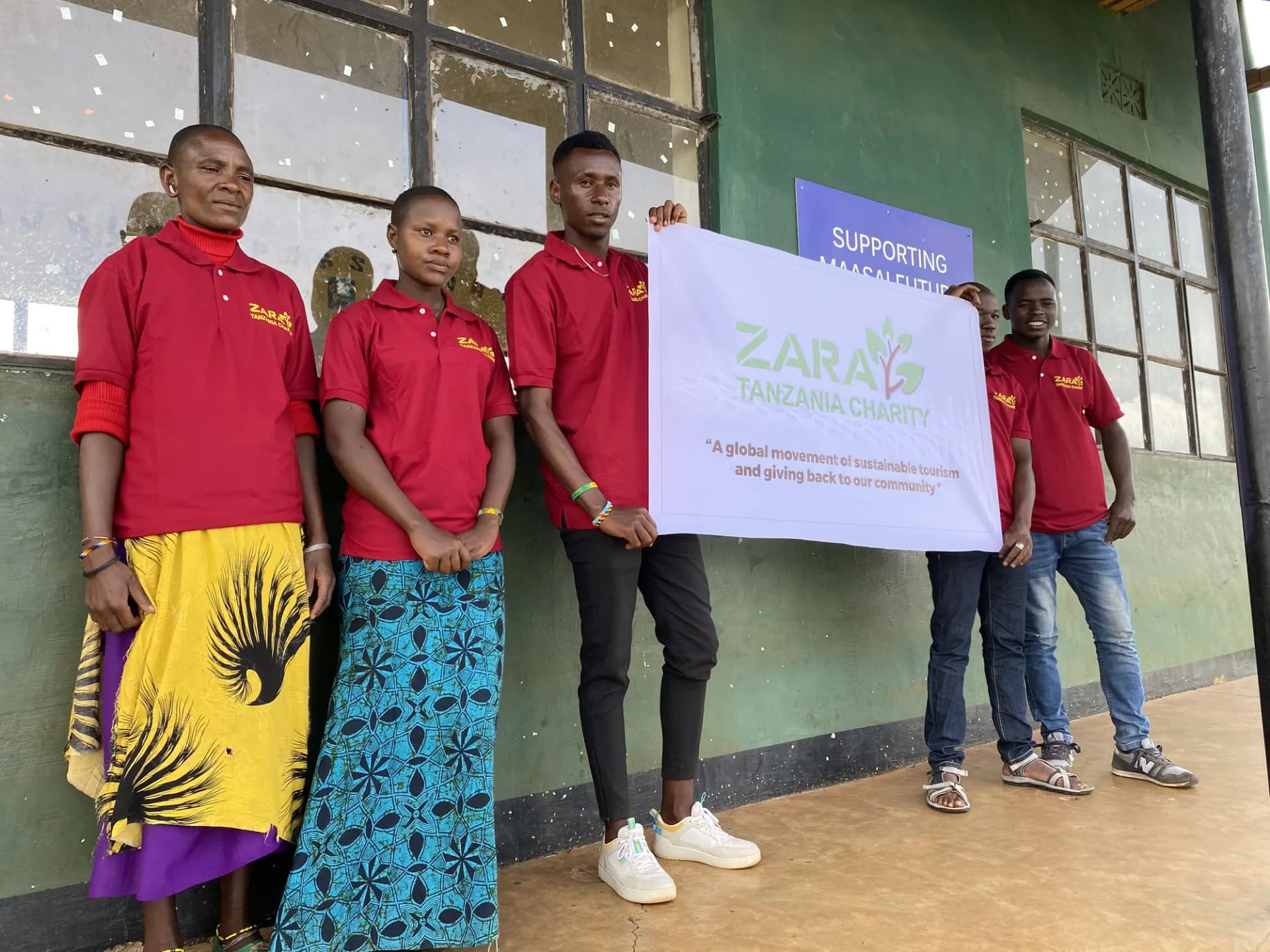 Young Maasai men and women in charity polo shirts and kanga wraps standing together outside the school.