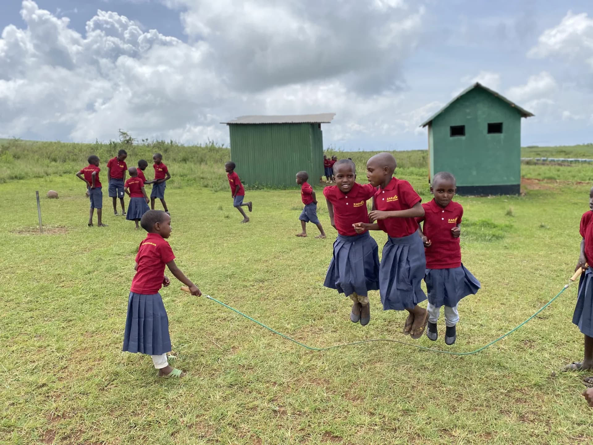 School children skipping a long rope together on the grass field, the green latrine blocks behind them.