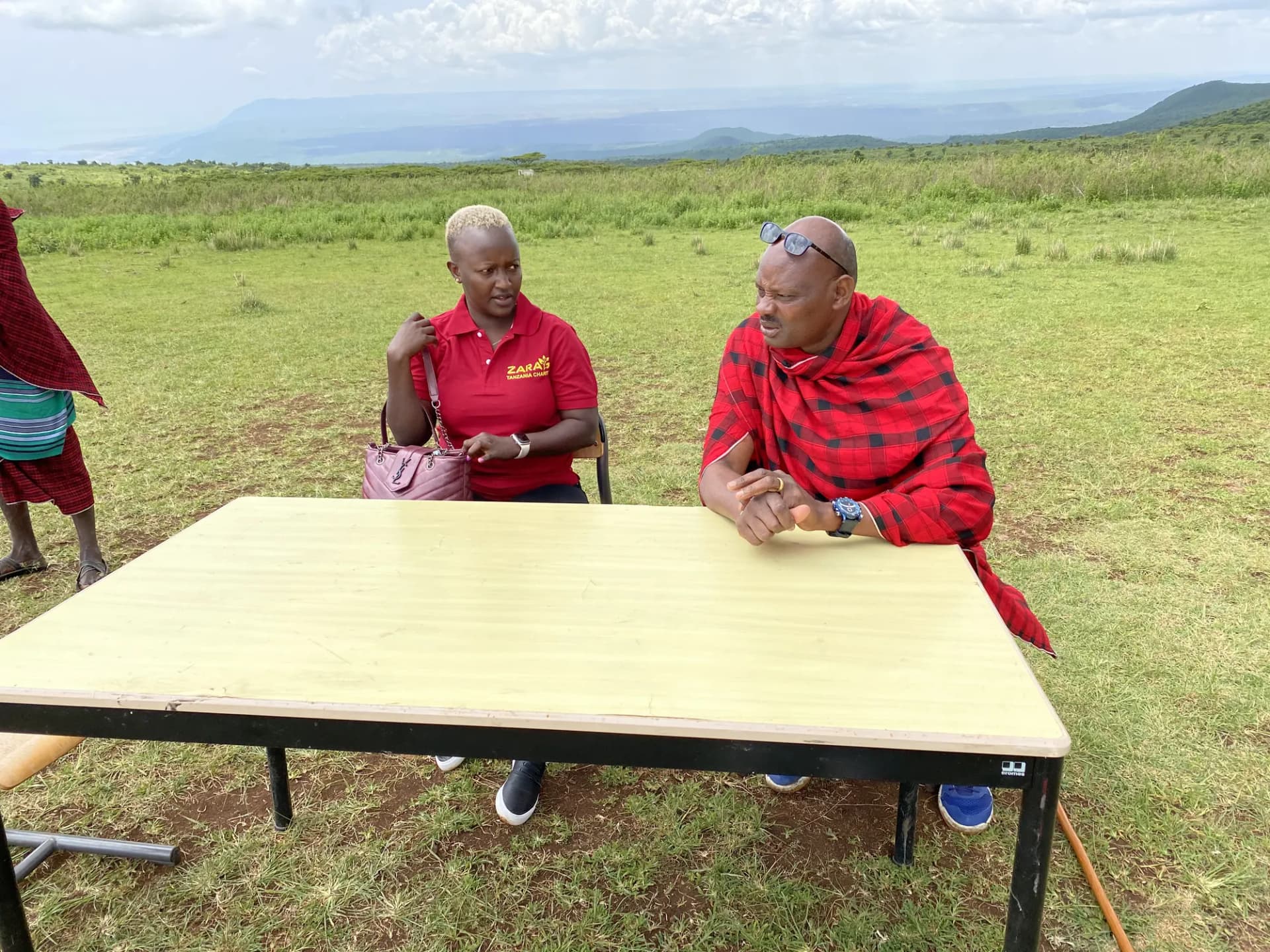 A charity fieldworker sitting at a table set up in open grassland, meeting a Maasai elder.