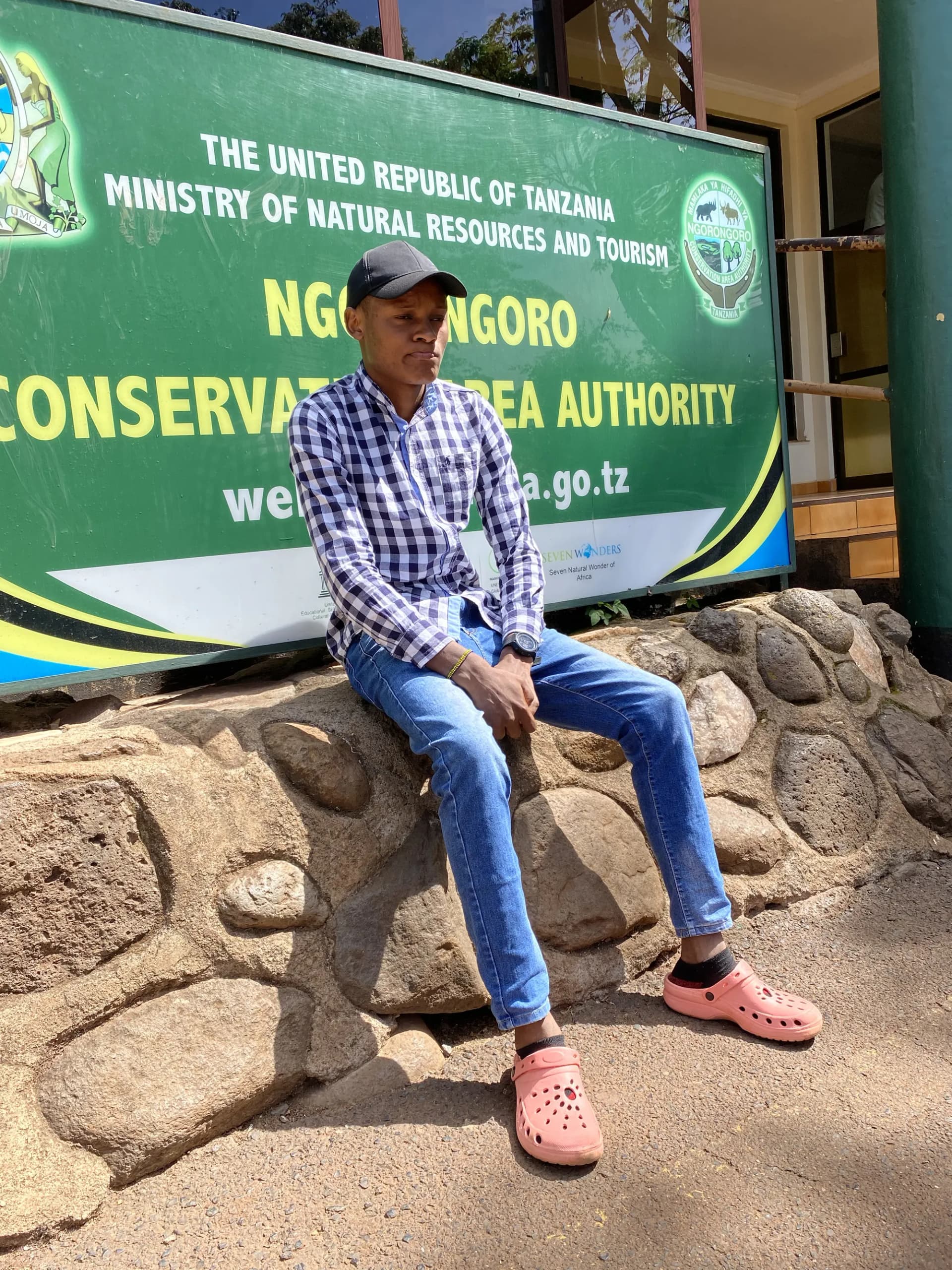 A staff member sitting on the stone wall beneath the Ngorongoro Conservation Area Authority sign.
