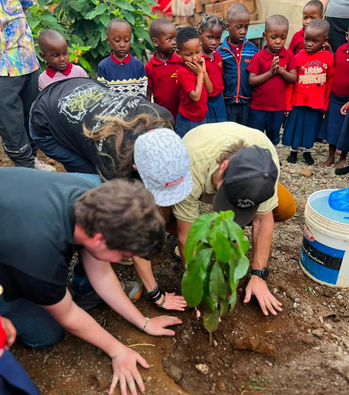 Visiting travellers kneeling in the soil to plant a tree seedling while school children in uniform watch from behind.