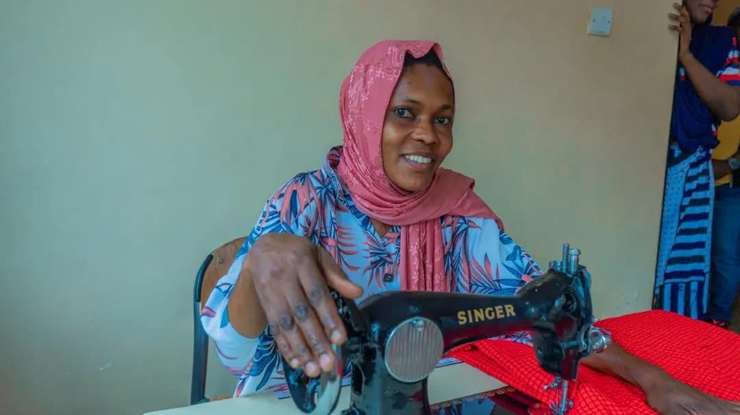 A woman in a pink headscarf smiling at her treadle sewing machine during tailoring training.