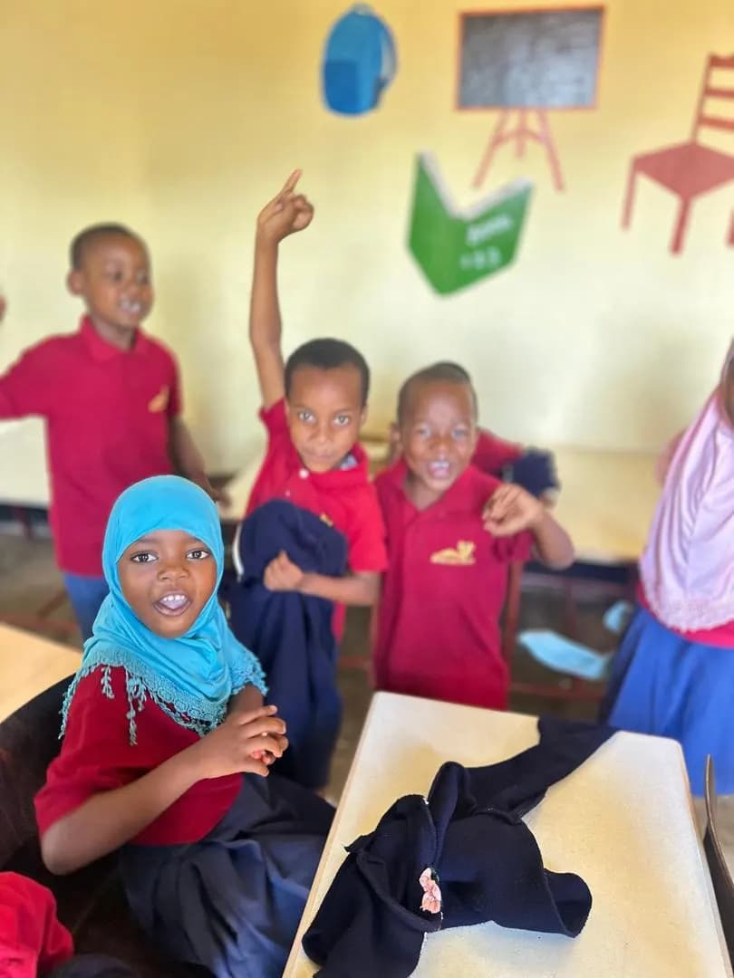 Young children in a classroom in red polo shirts, arms raised and calling out, one in a pale blue headscarf.