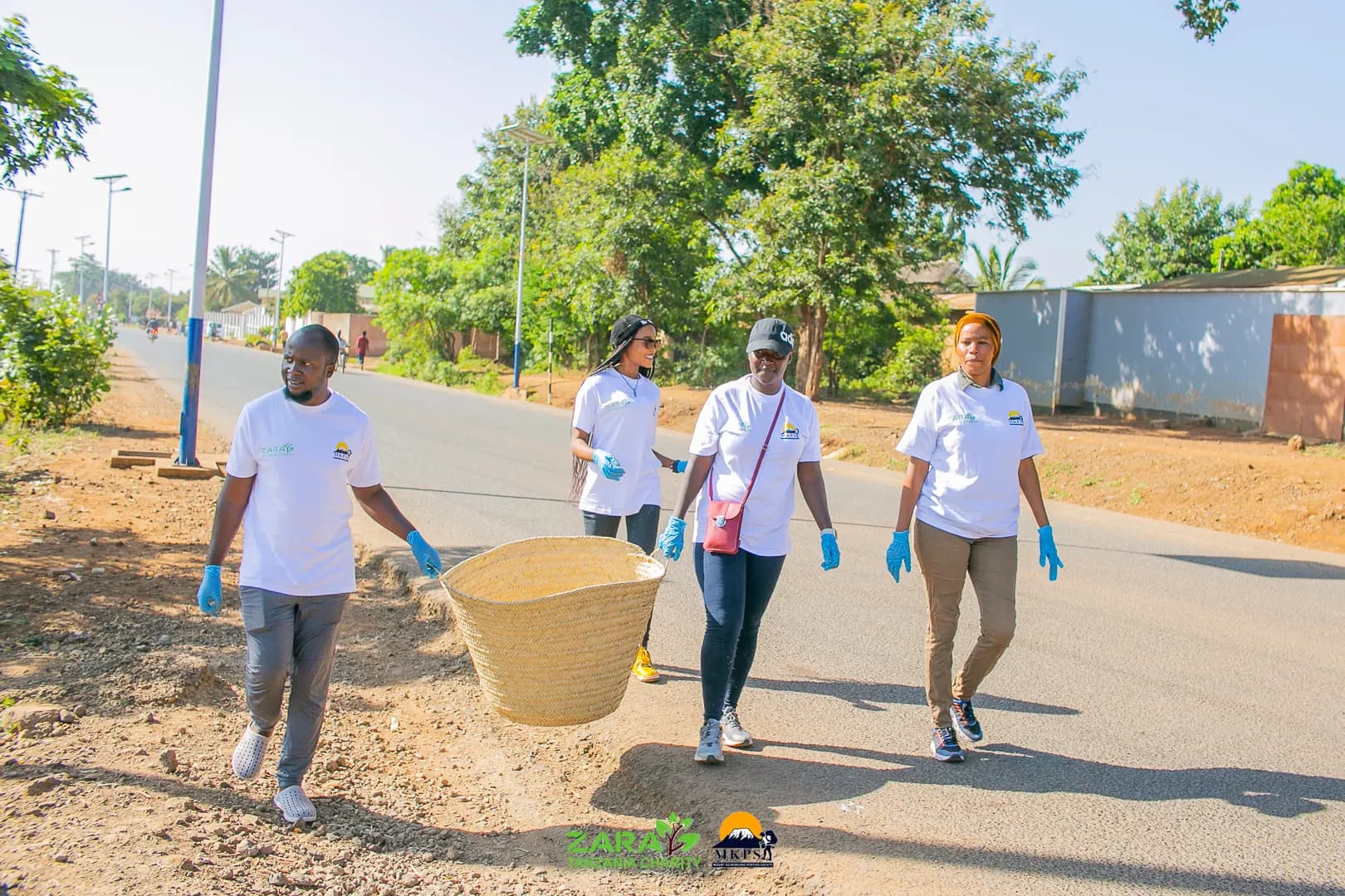 Five volunteers in white charity shirts walking down a tarmac road carrying a large woven basket between them during a roadside cleanup.