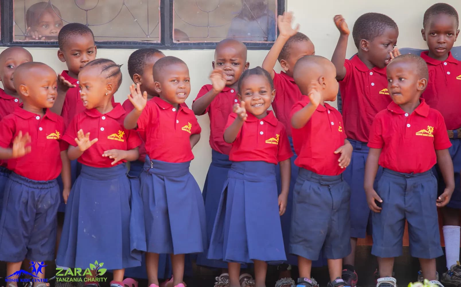 A close group of pre-school children in red polo shirts and navy uniforms waving and grinning inside their classroom at Moshi Kids Centre.