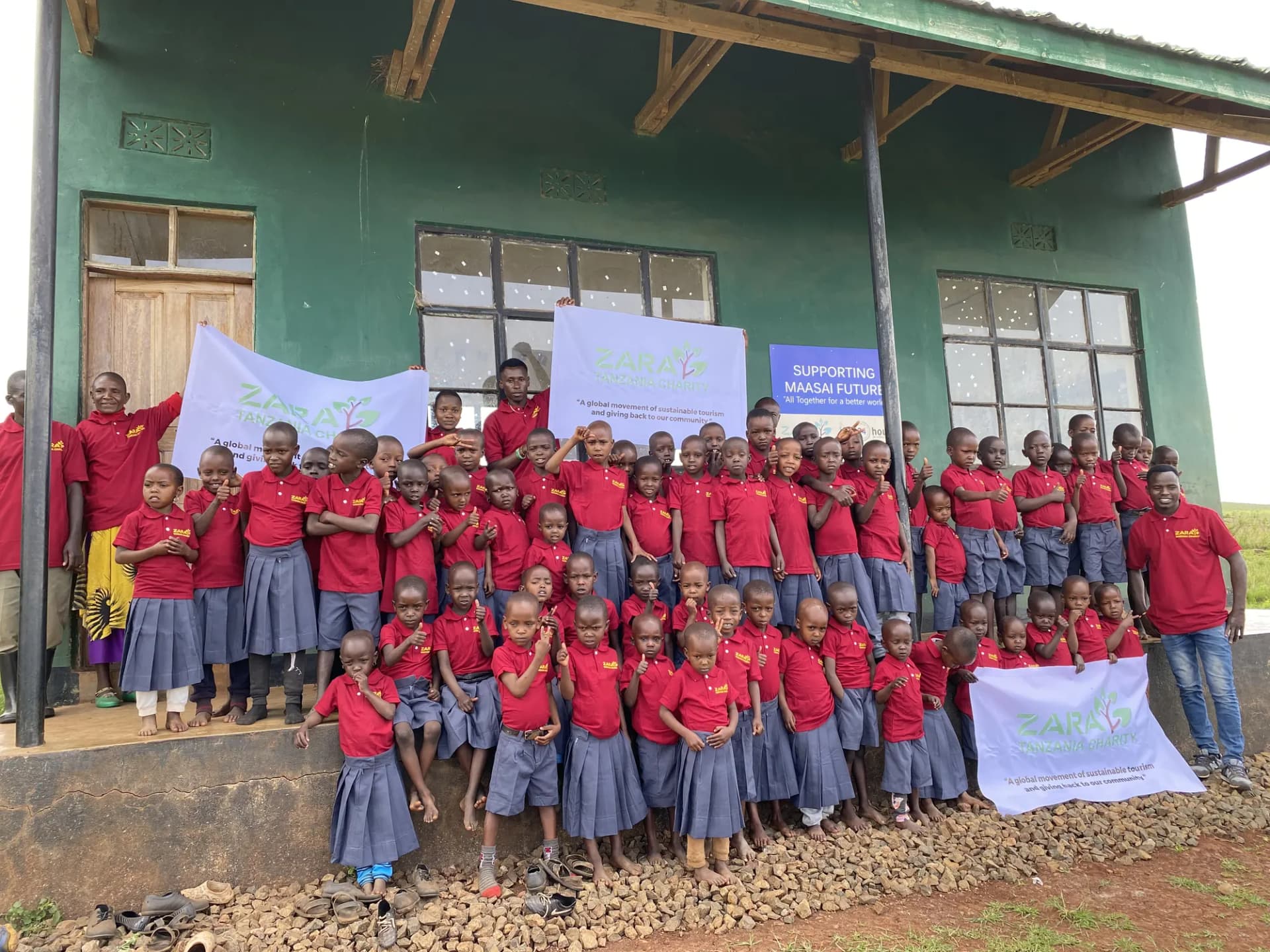 The whole Maasai Pre-School assembled in front of their green classroom block, holding the charity banner and a 'Supporting Maasai Future' sign.