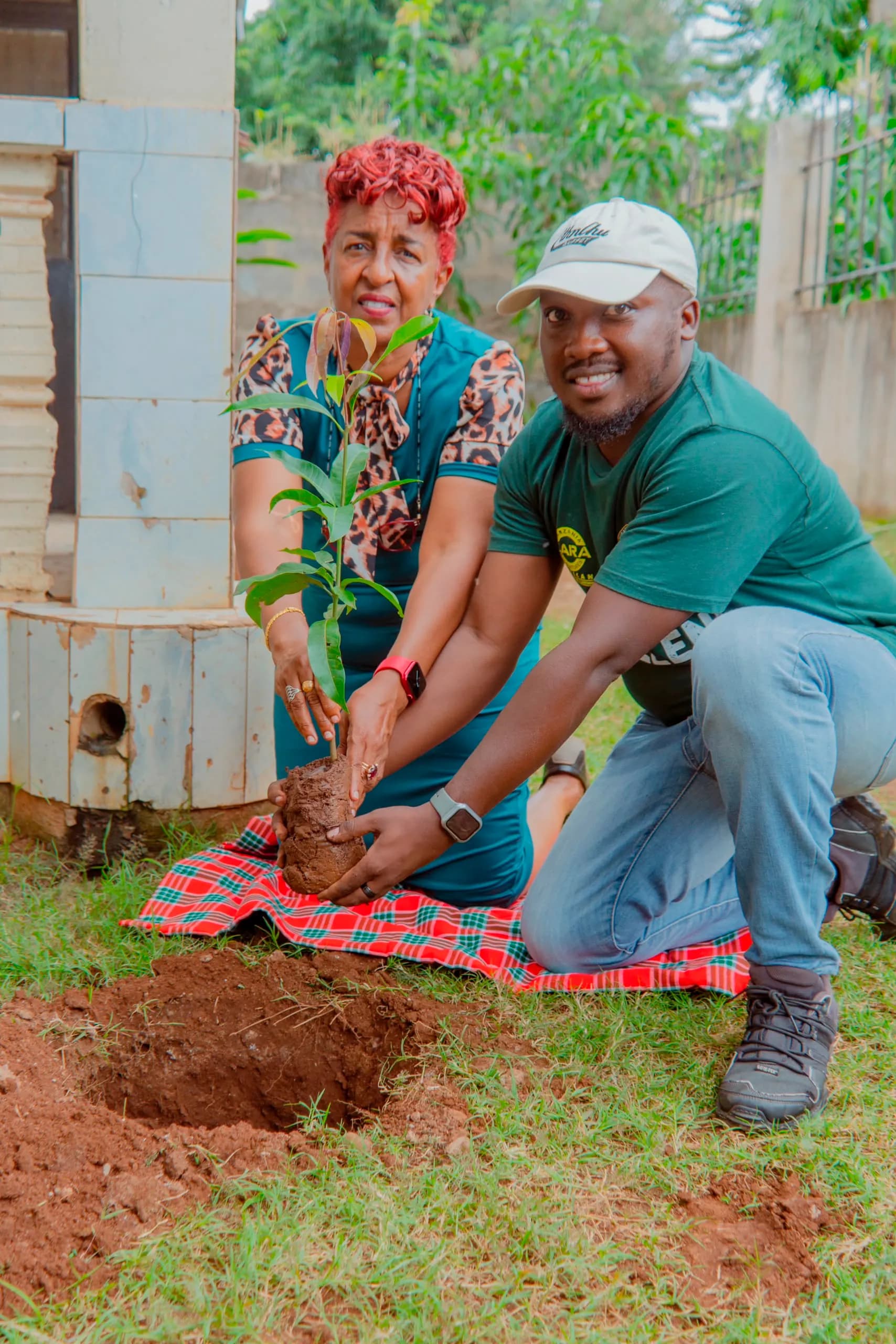 Two people kneeling on a Maasai blanket, easing a tree seedling out of its bag before planting it beside a water tank.