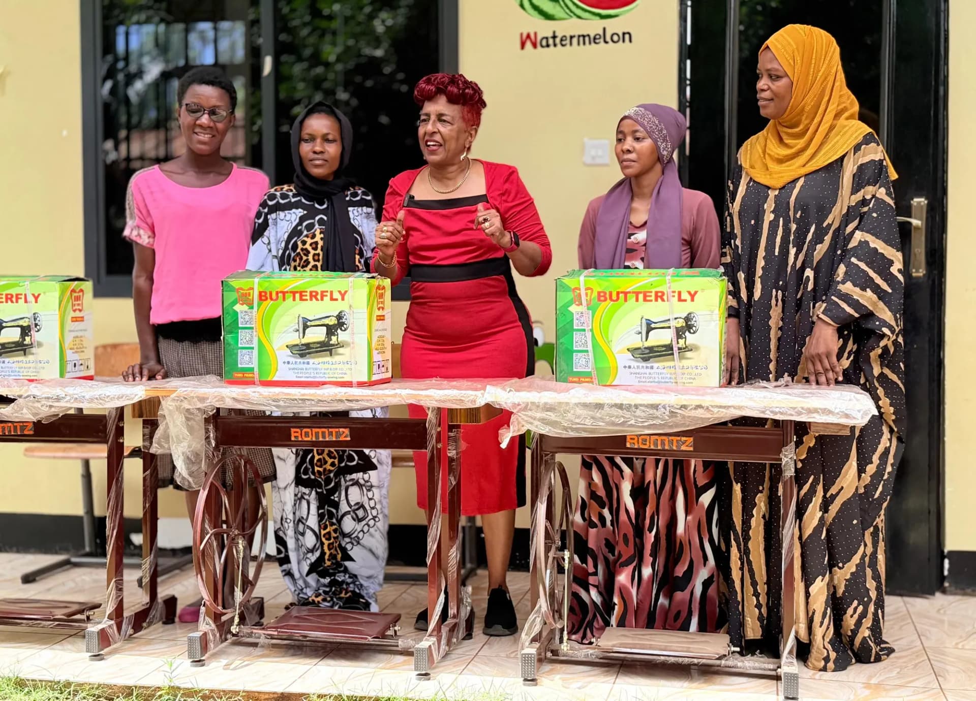 Zainab Ansell with women of a tailoring group at the handover of new treadle sewing machines, the boxed machines on the tables in front of them.