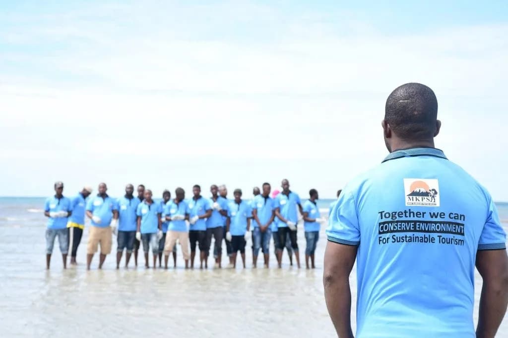 A Mount Kilimanjaro Porters Society volunteer watches from the foreground as a line of cleanup volunteers in blue shirts stands in the shallows along the Tanzanian coast. His shirt reads “Together we can conserve environment for sustainable tourism”.
